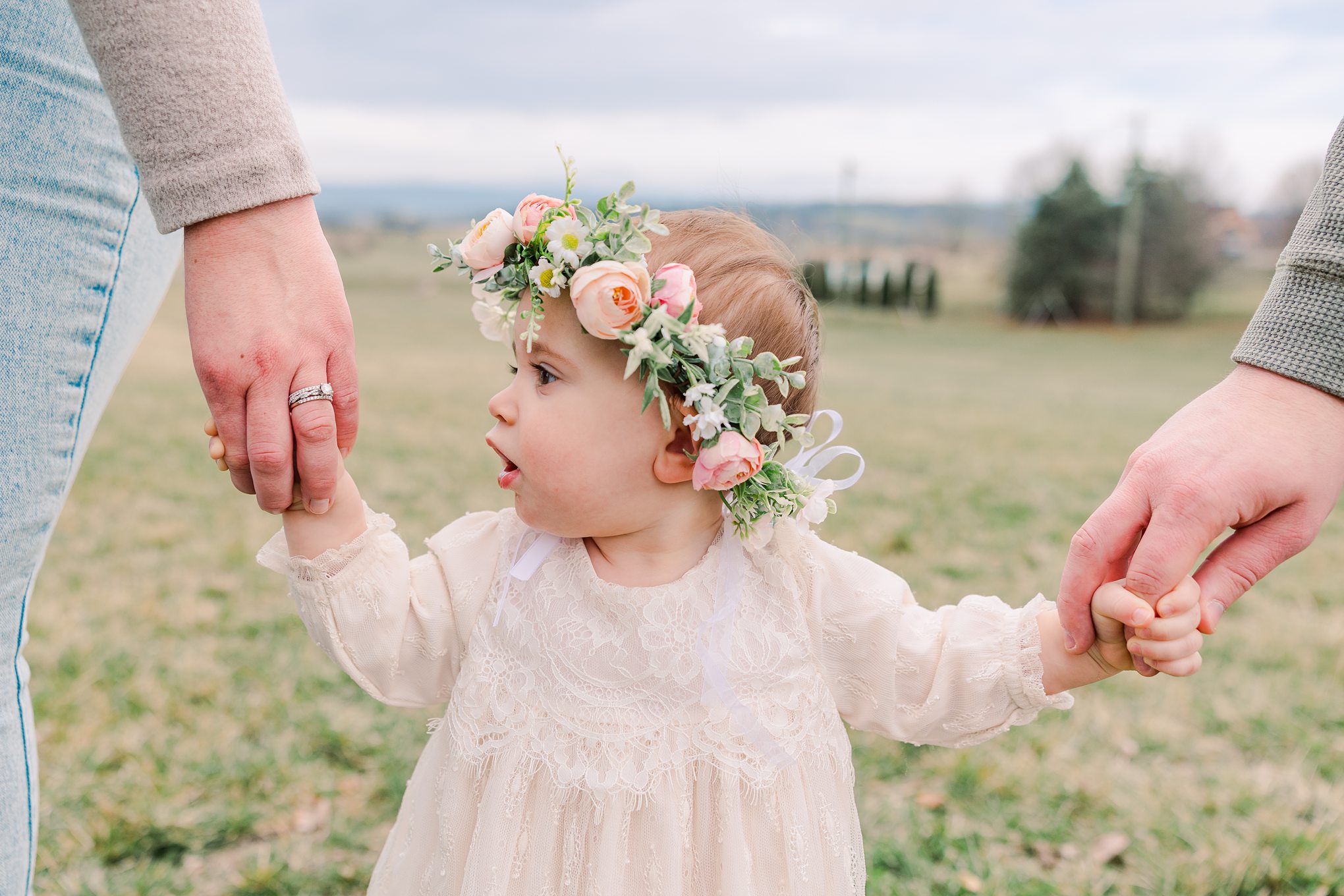 one year old in flower crown holding parents hands outside