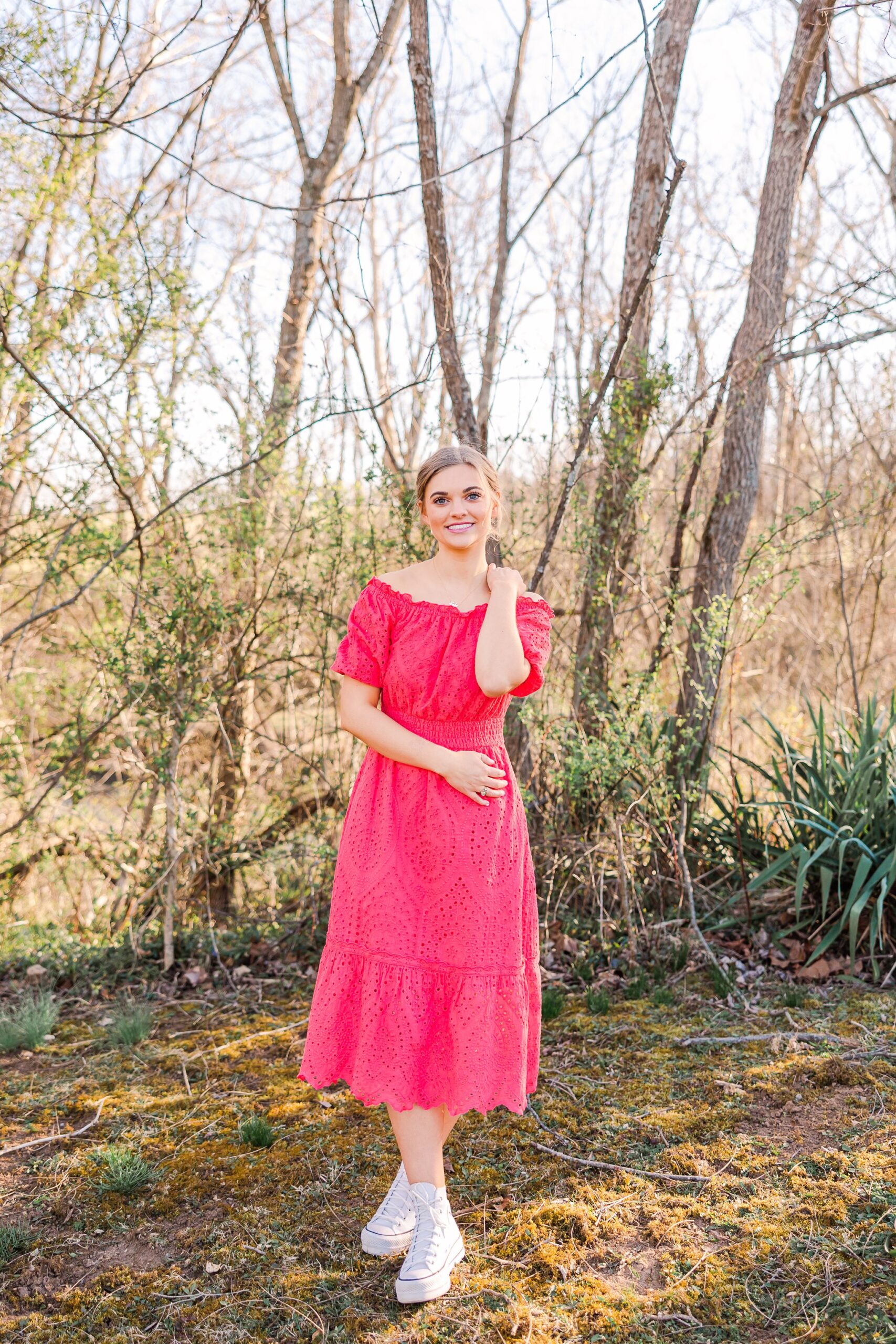 senior girl wearing pink off shoulder dress outside in woods touching her shoulder with one hand
