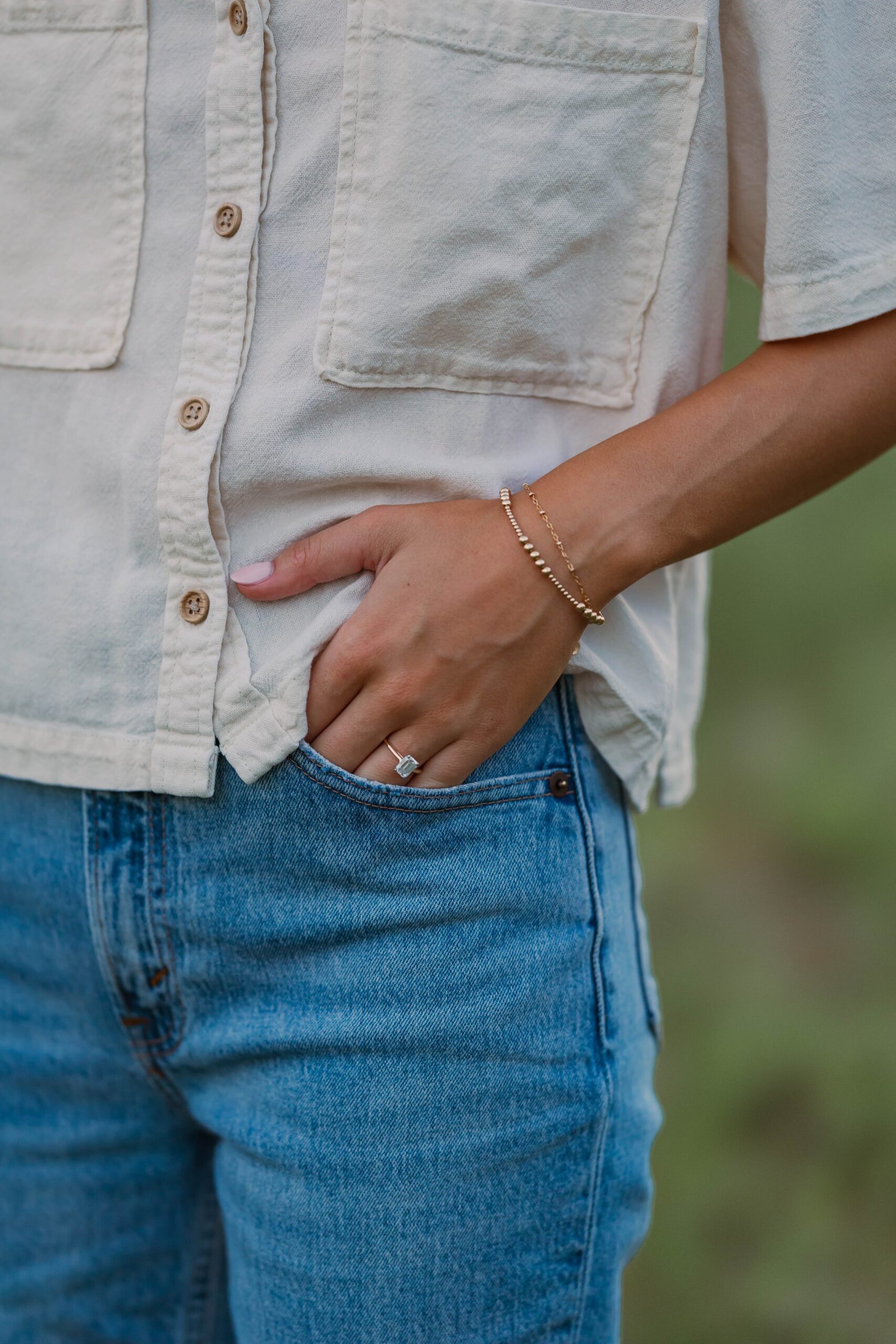 A close up shot of a woman's white shirt and jeans, with her hand styled in her pocket. On her hand is her engagement ring. 