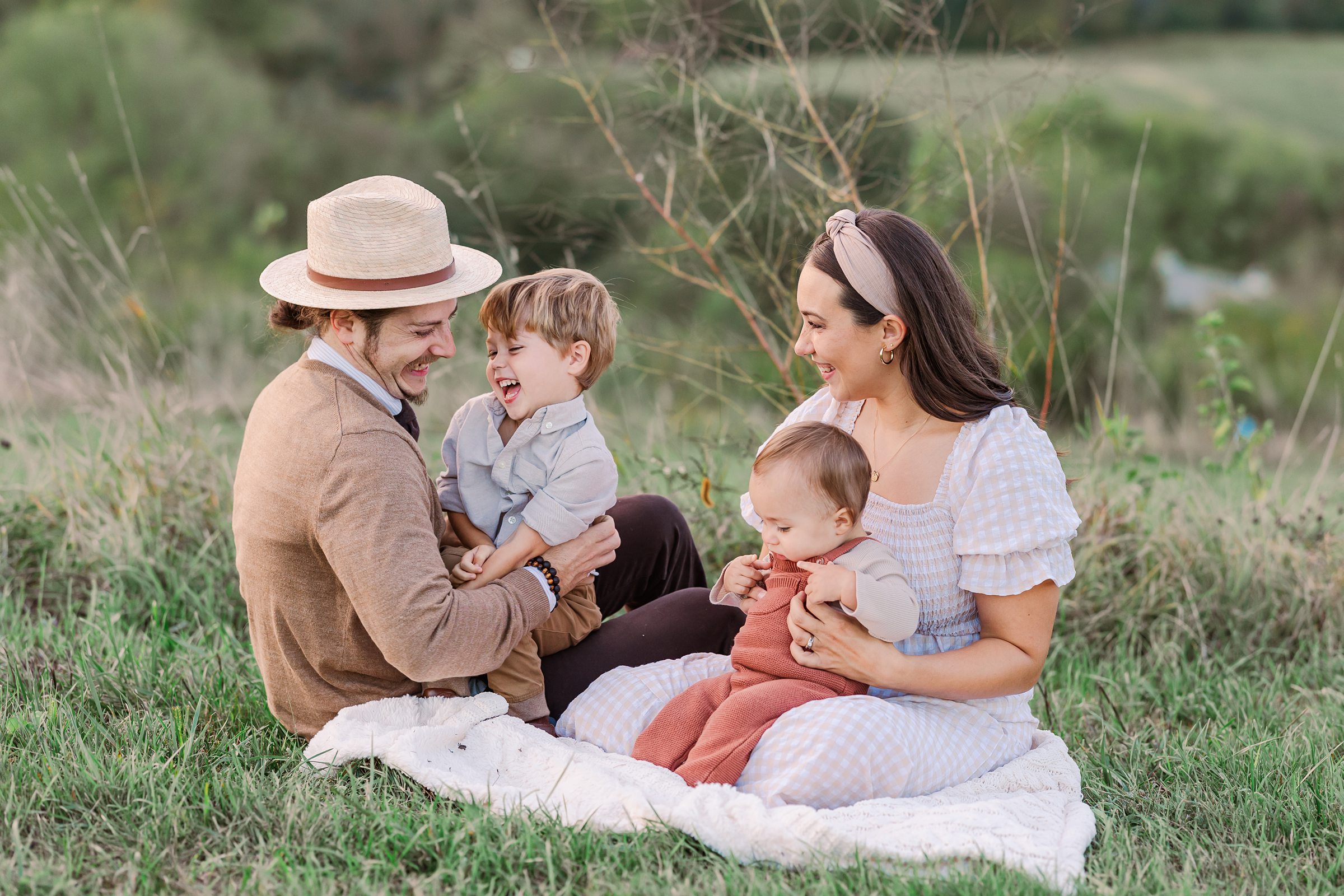 A young family sits on a white picnic blanket in a field of grass. The father is wearing a tan sweater and a flat brimmed hat, the mother is wearing a tan hairband and a tan and white checkered dress, one son is wearing a collared shirt and laughing and the baby is wearing rust-colored overalls. 