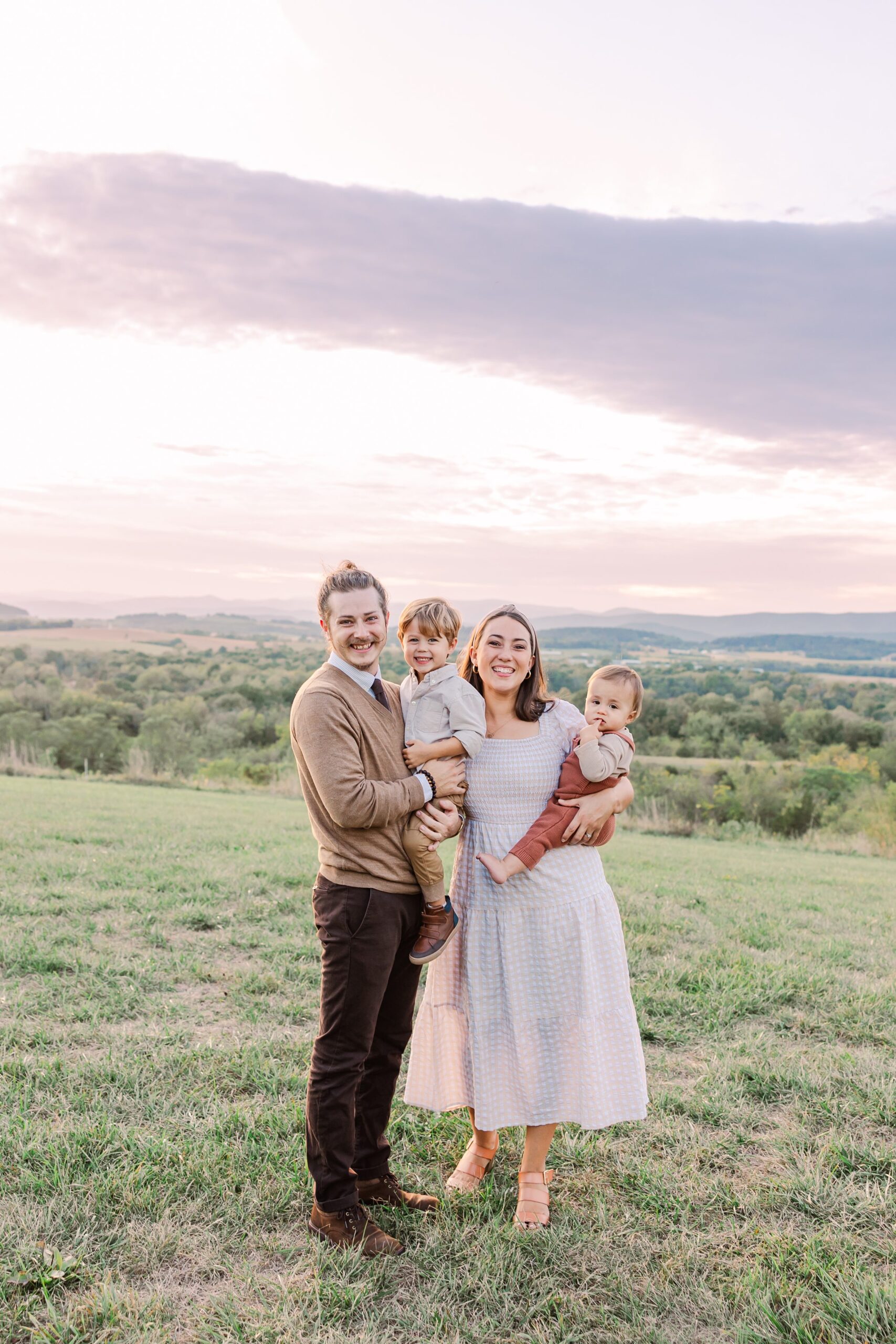 A young family with two young boys smile at the camera in a grassy field. The Blue Ridge mountains are in the background. 