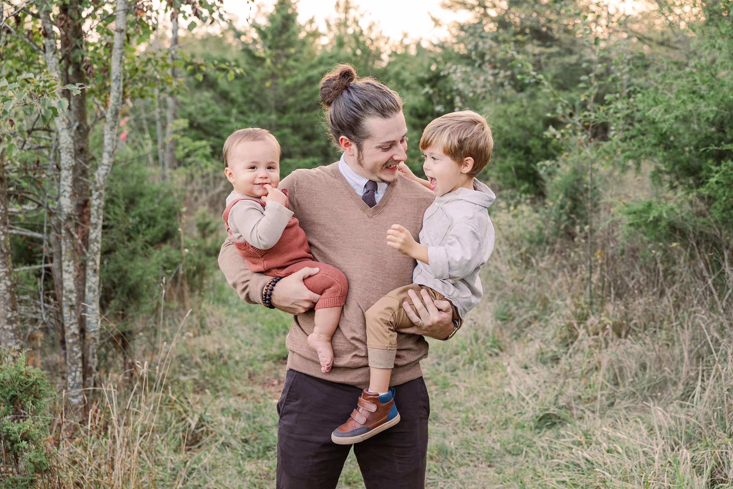 A young father is holding his two sons on his hips in a grassy field and smiling at his oldest child. The oldest child is smiling at his baby brother and the baby has a finger in his mouth. 