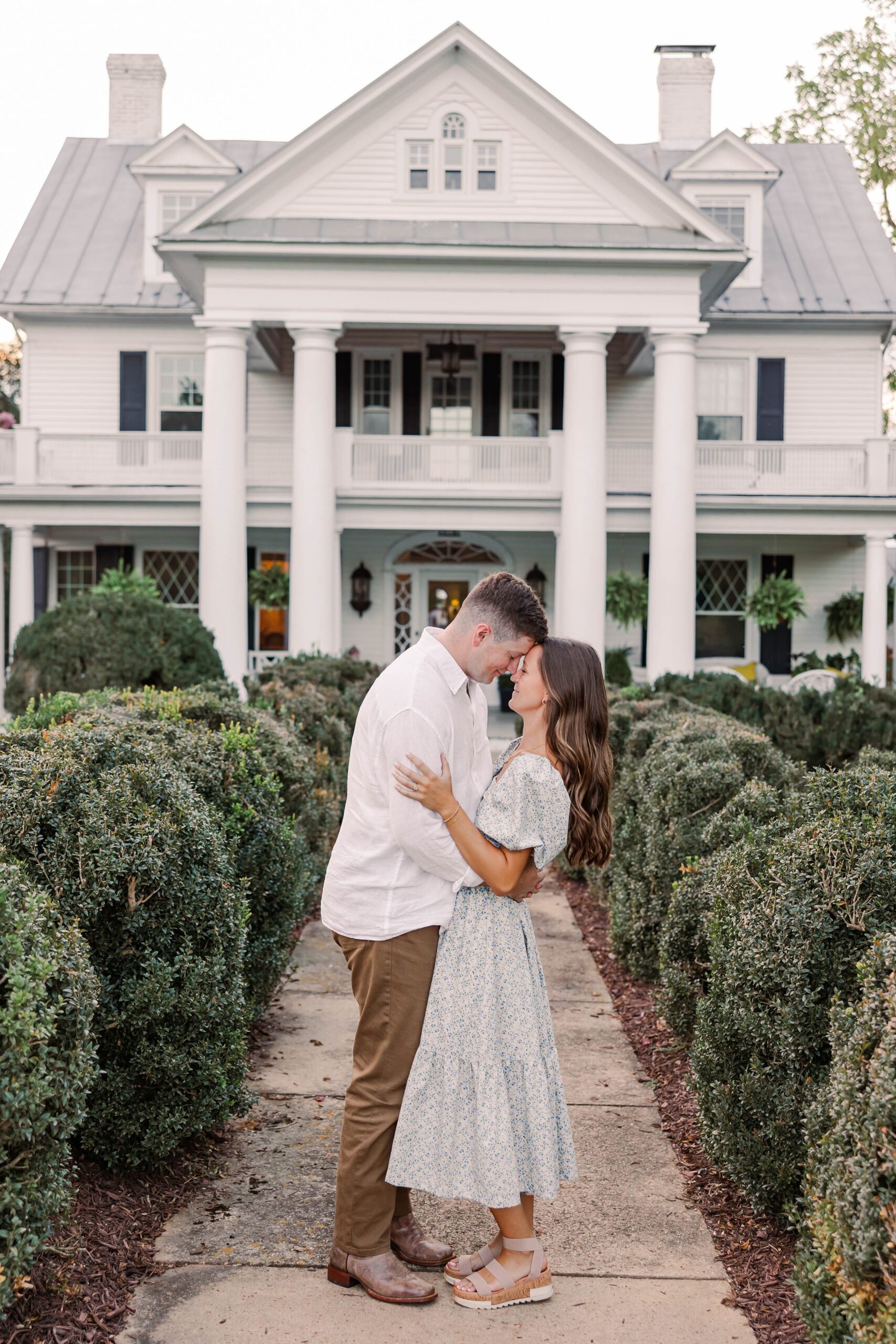 A young, engaged couple embrace in front of a large, white house with pillars. 